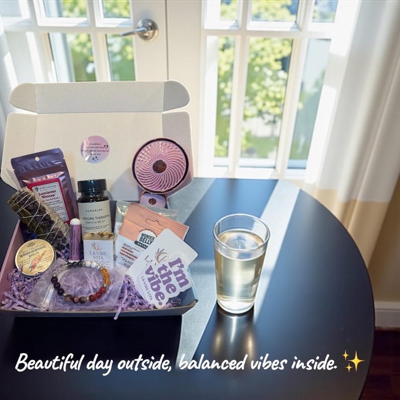 May include: A gift box filled with self-care items, including a small pink fan, a bracelet, and various beauty products. The box sits on a black table next to a glass of water. Text on the box reads "I'm the vibe."