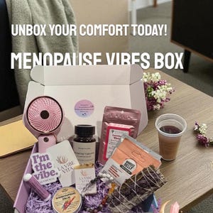 May include: A box labeled "MENOPAUSE VIBES BOX" filled with self-care items. Includes a pink fan, skincare products, tea, and a sage bundle. The box is on a table with the text "UNBOX YOUR COMFORT TODAY!" and "LA VIBE VITA" branding.