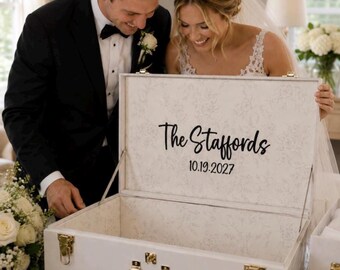 May include: A white rectangular wooden keepsake box with gold-toned metal latches and a handle. The interior lid features a subtle floral pattern and the black text The Staffords 10.19.2027. The box is open, revealing a spacious, empty interior.