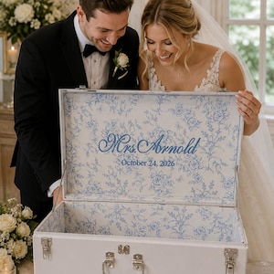 May include: A white rectangular storage trunk with silver metal latches and a handle. The interior is lined with a blue and white floral pattern. The text "Mrs. Arnold" and "October 24, 2026" is embroidered in blue thread on the inside lid.