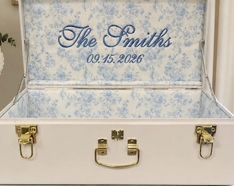 May include: A white storage trunk with gold-tone metal latches and a handle. The interior lid features blue floral fabric with the embroidered text The Smiths and the date 09.15.2026. The text Embroidered To last! is visible below the trunk.