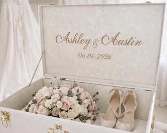 May include: A white rectangular keepsake box with gold-toned latches and handle. The interior lid features the text "Embroidered To last!" and "Ashley & Austin 06.06.2026". Inside the box sits a floral bouquet of light pink and white roses.