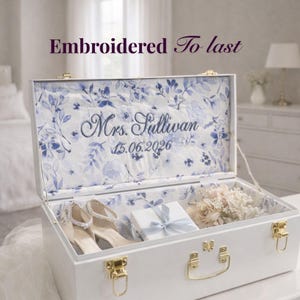 May include: White trunk with gold hardware, open to reveal a floral patterned interior. Inside are a pair of shoes, a gift box, and a bouquet. The text "Embroidered To last" and "Mrs. Sullivan 15.06.2026" are visible.
