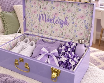 May include: A lavender storage trunk with gold-tone latches and a floral-lined interior featuring the name Marleigh. Inside are white sneakers, a lavender sweatshirt, a lavender and white hair bow, two purple and white pom-poms, and a bottle.