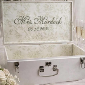 May include: A white rectangular storage chest with silver metal latches and a handle. The interior lid features a light green floral pattern with the embroidered text Mrs. Murdock and the date 06.12.2026 in a cursive font style.