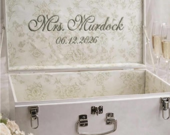 May include: A white rectangular storage chest with silver metal latches and a handle. The interior lid features a light green floral pattern with the embroidered text Mrs. Murdock and the date 06.12.2026 in a dark green script font.