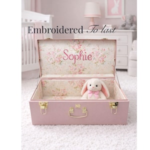 Pode incluir: Um baú rosa com ferragens douradas e interior floral. O nome "Sophie" é bordado no interior. Um coelhinho de pelúcia com um laço rosa está dentro. O texto "Embroidered To last" está acima.