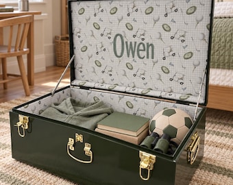 May include: A dark green metal storage trunk with gold-tone latches and handles. The interior lid features a white fabric lining with a repeating golf-themed pattern and the name Owen printed in the center. It contains a folded green garment, books, a soccer ball, and binoculars.