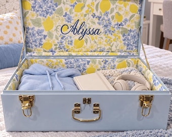 May include: A light blue storage trunk with gold-tone metal latches and a handle. The interior is lined with a floral pattern featuring yellow lemons and blue flowers, with the name Alyssa embroidered in dark blue script on the inside lid.