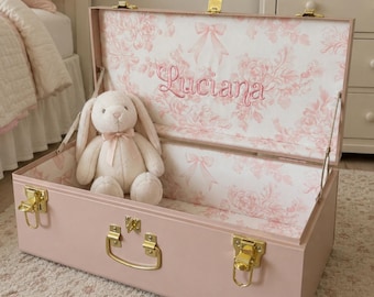 May include: A light pink storage trunk with gold-tone metal latches and a handle. The interior is lined with a white fabric featuring a pink floral and ribbon pattern, with the name Luciana embroidered in pink. A cream-colored plush rabbit sits inside.