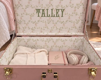 May include: A light pink storage trunk with gold-tone metal latches and a handle. The interior is lined with a white fabric featuring a green leaf and pink bow pattern. The name TALLEY is embroidered in green on the inside lid.