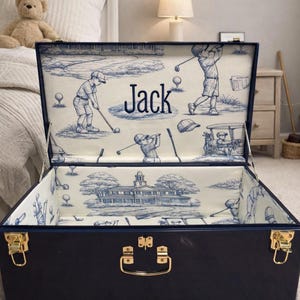 Puede incluir: Un ba&uacute;l de almacenamiento azul marino con herrajes dorados, abierto para revelar un forro interior con tem&aacute;tica de golf. El forro presenta un dise&ntilde;o toile de Jouy azul y blanco con escenas de golf y el nombre "Jack".