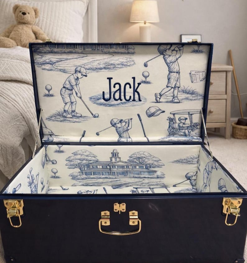 Puede incluir: Un ba&uacute;l de almacenamiento azul marino con herrajes dorados, abierto para revelar un forro interior con tem&aacute;tica de golf. El forro presenta un dise&ntilde;o toile de Jouy azul y blanco con escenas de golf y el nombre "Jack".