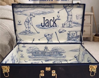 May include: A dark blue storage trunk with gold-toned metal latches and a handle. The interior is lined with cream fabric featuring a blue toile golf pattern and the name Jack embroidered in the center. The text Embroidered To last! is visible.
