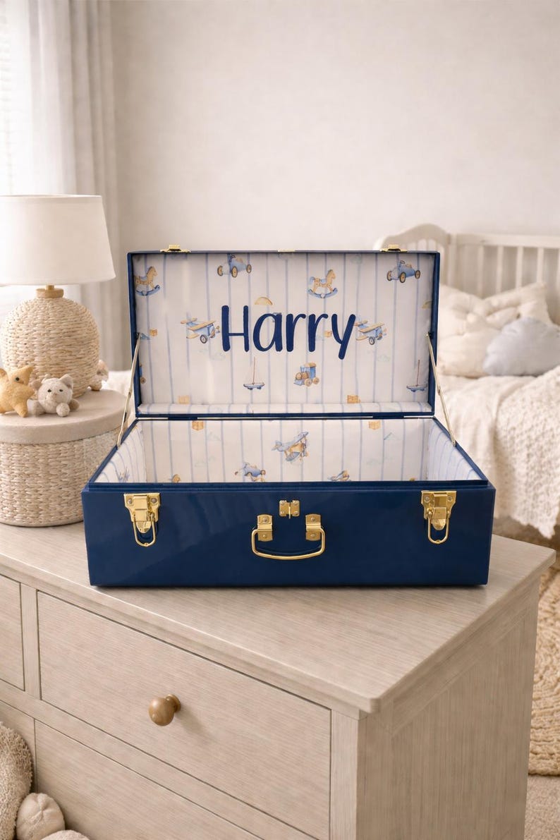 Puede incluir: Un ba&uacute;l de almacenamiento azul marino con herrajes dorados y el nombre "Harry" en escritura azul. El interior est&aacute; forrado con una tela blanca estampada con aviones de juguete, trenes y caballitos balanc&iacute;n. El ba&uacute;l est&aacute; abierto.