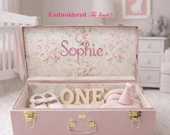 May include: A dusty pink storage trunk with gold-tone metal latches and a handle. The interior lid features a floral pattern and the embroidered name Sophie. Inside, wooden letters spell ONE, accompanied by white baby shoes and a pink party hat.