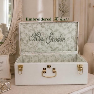 May include: A white rectangular storage chest with gold-toned metal latches and a handle. The interior lid features a light green floral pattern and the embroidered text "Mrs. Gooden" in a cursive script, with "Embroidered To last!" above.