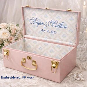 May include: A rectangular pink storage box with gold-tone metal latches and a handle. The interior is lined with a white fabric featuring a blue floral pattern. The lid interior displays the blue embroidered text Megan & Matthew May 30, 2026.