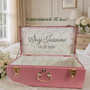 May include: A pink wooden keepsake box with gold-tone metal latches and a handle. The interior lid features a white fabric lining with a green vine pattern, the embroidered name Ivy Jeanne, and the date 06.22.2026 in grey thread.