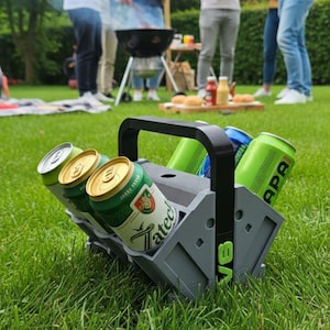 Może przedstawiać: Szary uchwyt na piwo z kilkoma puszkami piwa, w tym marki Zatec i APA, stoi na trawniku. Uchwyt ma czarny uchwyt. W tle widać ludzi i grill.