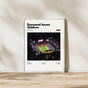 May include: A framed print featuring an aerial view of Raymond James Stadium in Tampa Bay, Florida. The stadium is lit up at night, with the field and seating areas visible. Text at the top reads "Raymond James Stadium" and "TAMPA BAY". Additional text includes coordinates, capacity, and the year opened.
