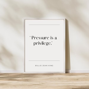 May include: A framed print with the quote "Pressure is a privilege." in black text on a white background. The quote is attributed to Billie Jean King. The frame is white, and the print is displayed against a light-colored wall and wooden floor.