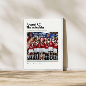 Op de afbeelding: Een ingelijste poster ter herdenking van de Premier League-overwinning van Arsenal F.C. in 2003/2004, met de titel "The Invincibles". De afbeelding toont het team in rood en wit, met de trofee. De poster bevat ook de tekst "Arsenal F.C.".