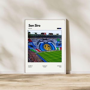 May include: A framed print featuring the San Siro stadium in Milan. The image shows a vibrant crowd with a large banner displaying a team logo. Text at the top reads "San Siro, Milan." Additional text includes coordinates, capacity, and the year 1926.