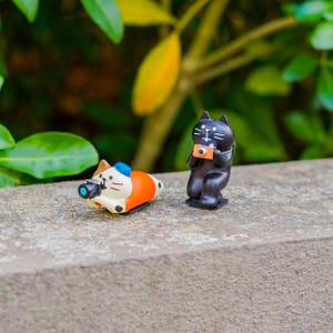 May include: Two cat figurines, one orange and white with a blue hat and the other black, both holding cameras. The figurines are on a concrete surface with a blurred green background of leaves and foliage.