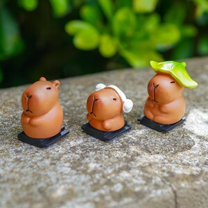 Può includere: Tre piccole statuette di capibara marroni su basi rettangolari nere. La figura a sinistra è semplice, quella centrale indossa cuffie bianche e quella a destra ha una foglia verde brillante sulla testa. Tutte hanno gli occhi chiusi.