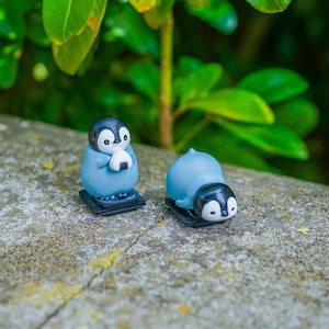 Puede incluir: Dos pequeñas figuras de pingüinos con cabezas negras y cuerpos azul claro. Un pingüino está de pie sosteniendo una bola de arroz triangular blanca con una tira de alga negra, mientras que el otro pingüino está tumbado boca abajo sobre una base negra.