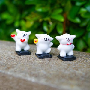 May include: Three small white animal figurines with simple black facial features. The left figure holds a red heart, the center figure holds a yellow flower, and the right figure wears a thin red neck ribbon. Each stands on a small black base.