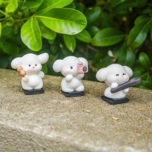 Puede incluir: Tres figuras de perros de dibujos animados blancos sobre bases negras. Uno sostiene un pincho de comida, otro una cámara rosa y el tercero un monopatín gris. Las figuras tienen mejillas rosadas, sobre un fondo de hojas verdes.