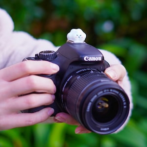 May include: A black Canon DSLR camera with a lens attached. A small, white cloud-shaped accessory with a smiling face is on top of the camera. The camera is held by a person.