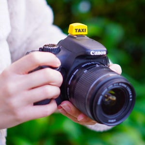 Può includere: Una fotocamera DSLR Canon nera con obiettivo zoom è tenuta in entrambe le mani. Un accessorio giallo per il tetto del taxi con la scritta "TAXI" è attaccato alla parte superiore della fotocamera. Lo sfondo è verde sfocato.