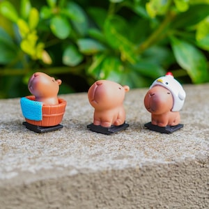 May include: Three miniature brown capybara ornaments. One is in a brown tub with a blue towel, another stands alone, and the third wears a white chicken hat with a red comb. Each figurine sits on a small black base.