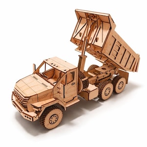 Puede incluir: Maqueta de madera de un camión volquete con la caja levantada. El camión está hecho de madera de color claro y presenta una construcción detallada, incluyendo la cabina, las ruedas y la caja. La palabra "DECKERY" es visible en la parte delantera.
