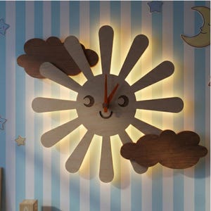 May include: Wooden sun-shaped wall clock with a smiling face and orange clock hands. The clock has a light-up feature and is surrounded by wooden cloud accents. The clock is mounted on a blue and white striped wall.