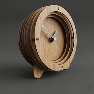Puede incluir: Un reloj de madera en forma de barril con un acabado marrón claro. El reloj presenta un diseño en capas con una esfera circular, manecillas plateadas y pequeños marcadores cuadrados. Se apoya sobre dos patas curvas.