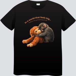May include: Black t-shirt featuring a graphic of two monkeys hugging. One is a plush toy, the other a real monkey. The text "In a world that feels big... hold on to something small." is printed above the image in orange.