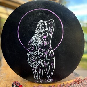 May include: A circular, black drum featuring a white illustration of a woman and a wolf within a purple circle. The woman has long hair and is decorated with floral and geometric designs. A wooden drumstick with a patterned head is below.