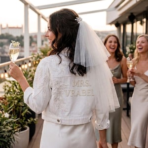 May include: A white denim jacket featuring intricate floral embroidery on the shoulder and the text MRS. VRABEL spelled out in pearl-like beads on the back. The jacket is worn with a short, layered white tulle veil attached to the hair.