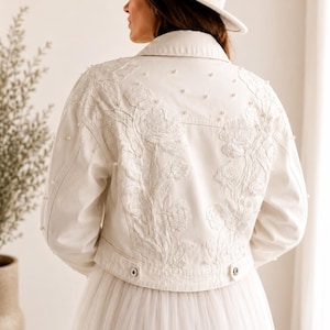 Könnte beinhalten: Eine weiße Jeansjacke mit floraler Stickerei und Perlenakzenten. Die Jacke hat einen klassischen Kragen, Knöpfe und lange Ärmel. Das Model trägt einen weißen Hut mit Perlenband und einen weißen Tüllrock. Ein Braut- oder Festtags-Outfit.