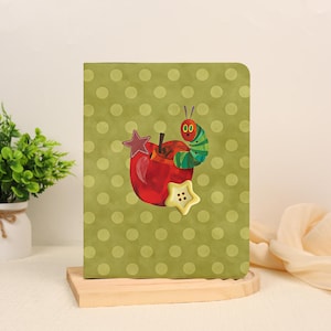 May include: A rectangular notebook with a light olive green cover featuring a repeating pattern of darker green polka dots. The center displays a red apple illustration, a green caterpillar, a maroon star-shaped patch, and a cream star-shaped button.