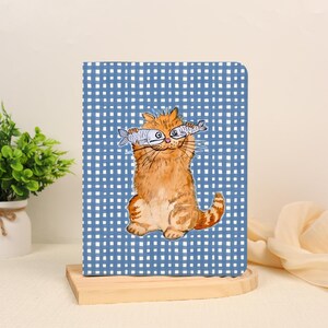 May include: A rectangular notebook with a blue and white checkered pattern cover. The center features an illustration of a fluffy orange cat holding two small silver fish over its eyes. The cat has a smiling expression and a long, striped tail.