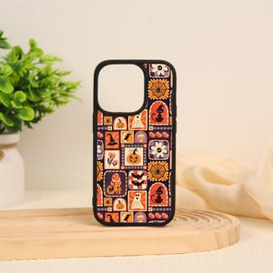 May include: A black phone case featuring a repeating grid pattern of Halloween-themed illustrations including orange pumpkins, black cats in witch hats, white ghosts, spider webs, bats, mushrooms, and flowers on a dark background.