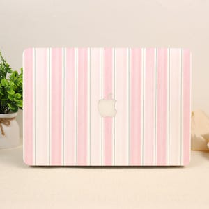 Peut inclure: Une coque rigide rectangulaire pour ordinateur portable avec un motif à rayures verticales alternant des tons rose clair, blanc et crème. Le centre de la coque présente une découpe pour le logo Apple, révélant la surface métallique en dessous.