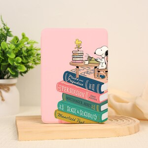 May include: A light pink e-reader case featuring a graphic illustration of Snoopy and Woodstock sitting at a table on a stack of Jane Austen books. The titles Pride and Prejudice, Persuasion, Northanger Abbey, Sense and Sensibility, and Mansfield Park are visible.