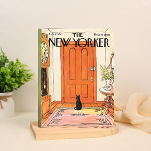 Może przedstawiać: Prostokątny notatnik z ilustracją z okładki magazynu The New Yorker z 4 lutego 1974 roku. Grafika przedstawia czarnego kota siedzącego na różowym dywaniku przed pomarańczowymi drzwiami, zegar szafkowy oraz stolik z bukietem kwiatów.