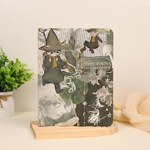 Può includere: Un taccuino rettangolare con un design a collage che mostra un personaggio dei cartoni animati con un cappello a tesa larga, note musicali, fiori e il testo SITTING QUIETLY, DOING NOTHING. La copertina ha toni verdi e marroni con varie illustrazioni.